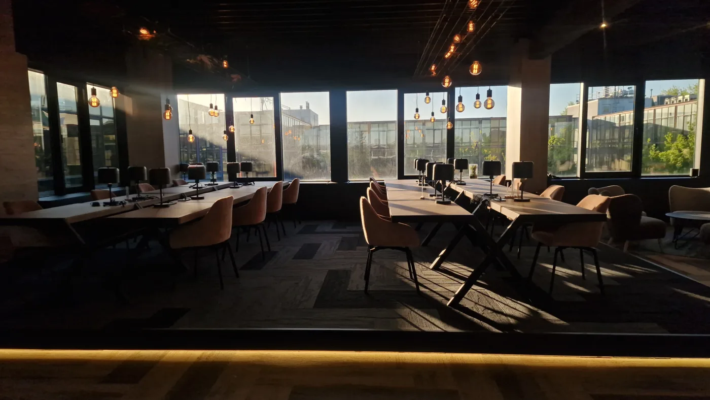 Dimly lit conference room with long white tables, beige chairs, and hanging Edison bulb lights along large windows.