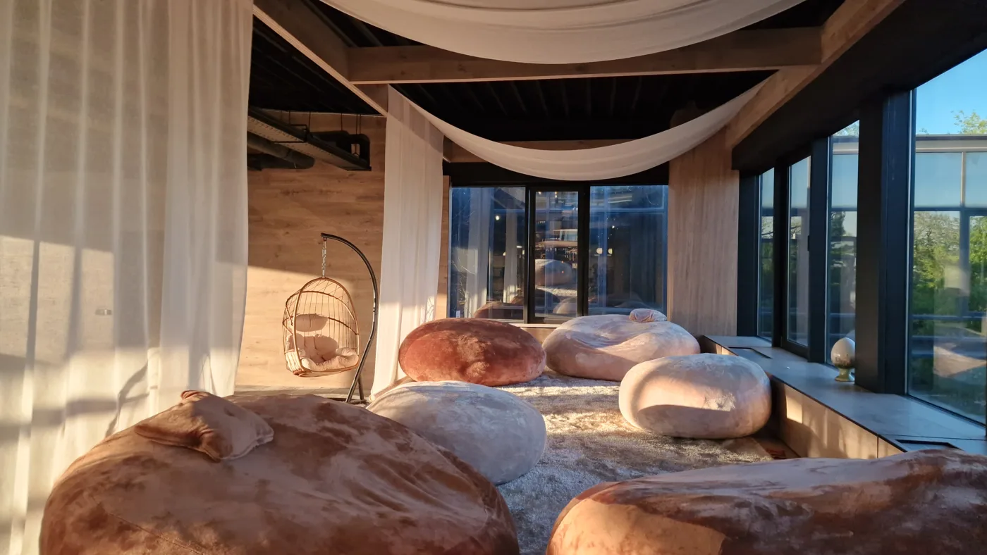 A sunlit lounge area with oversized brown beanbags, a hanging wicker chair, sheer curtains, and large windows along one wall.