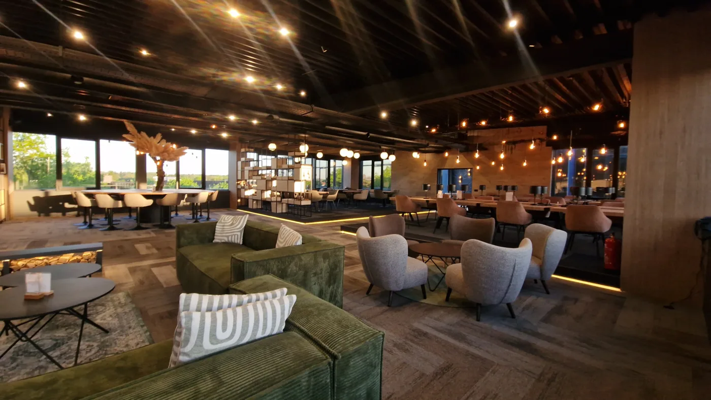 Spacious lounge with green couches, striped pillows, and round tables under warm hanging lights by large windows at dusk