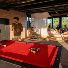 Man in an olive T-shirt takes a pool shot at a red billiards table in a modern lounge with hanging wicker chairs and large windows behind him.