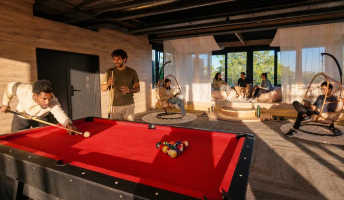 Man in an olive T-shirt takes a pool shot at a red billiards table in a modern lounge with hanging wicker chairs and large windows behind him.