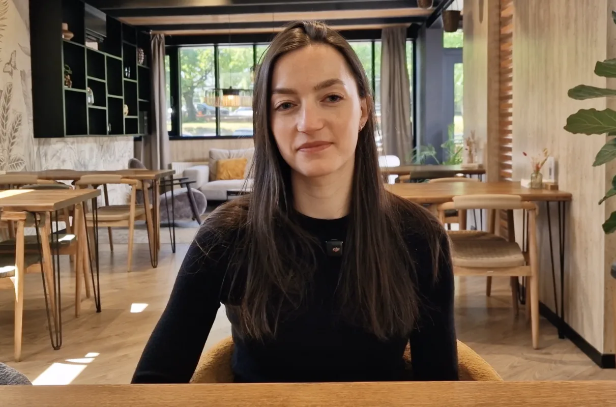 Woman with long brown hair in a black top sitting at a wooden table in a bright cafe interior with plants and large windows behind her, looking at the camera; neutral expression