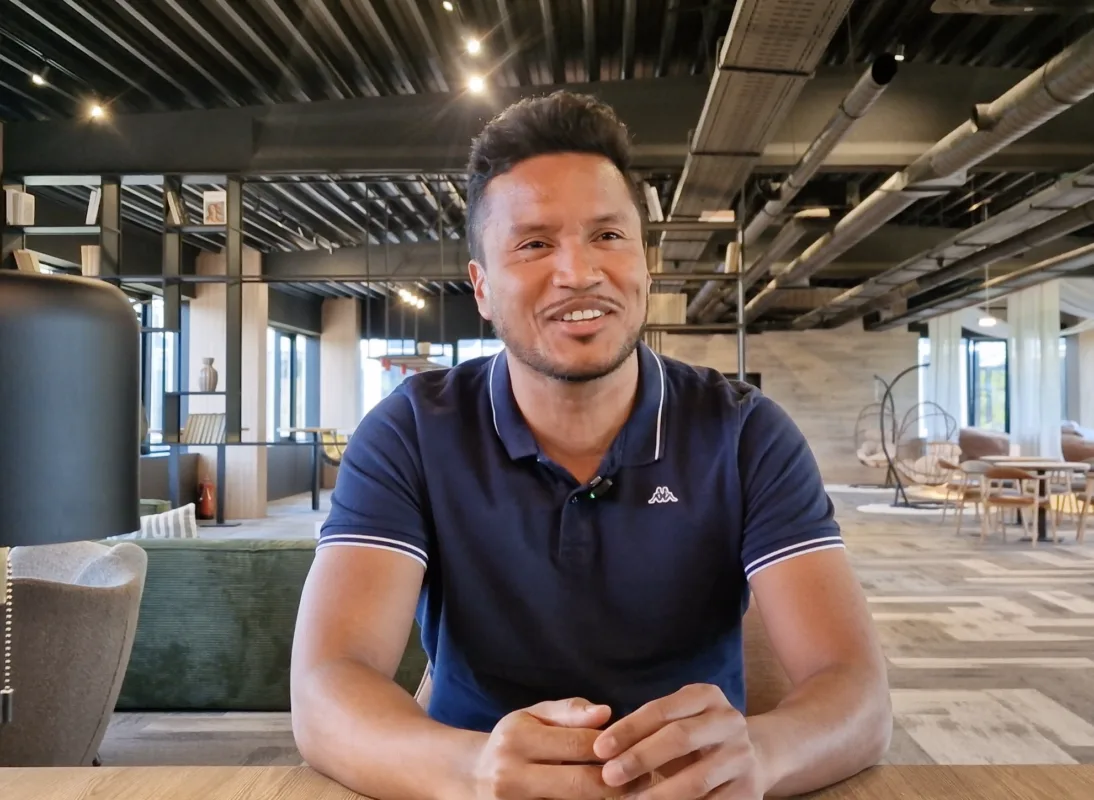 Smiling man with short dark hair in a navy polo sits at a wooden table in a modern, open-plan cafe.